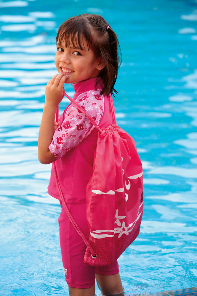 Young girl dressed in pink stood by a pool with a pink drawstring bag on her back.